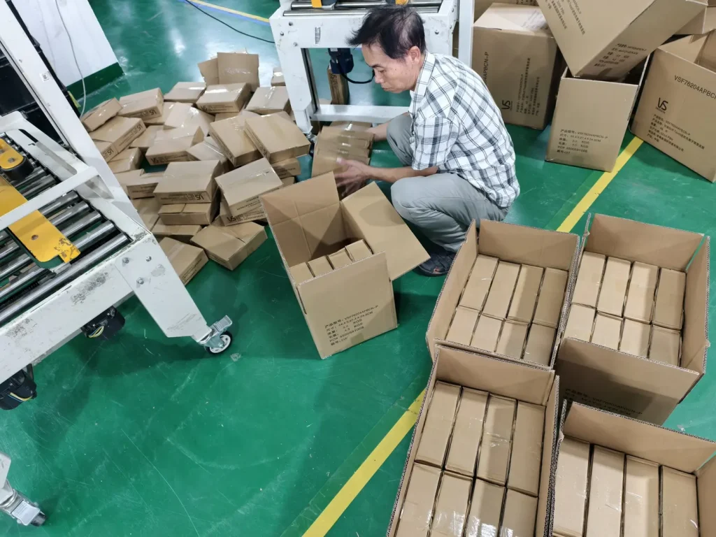 Worker packing small boxes into cartons beside stacks of other shipping boxes.