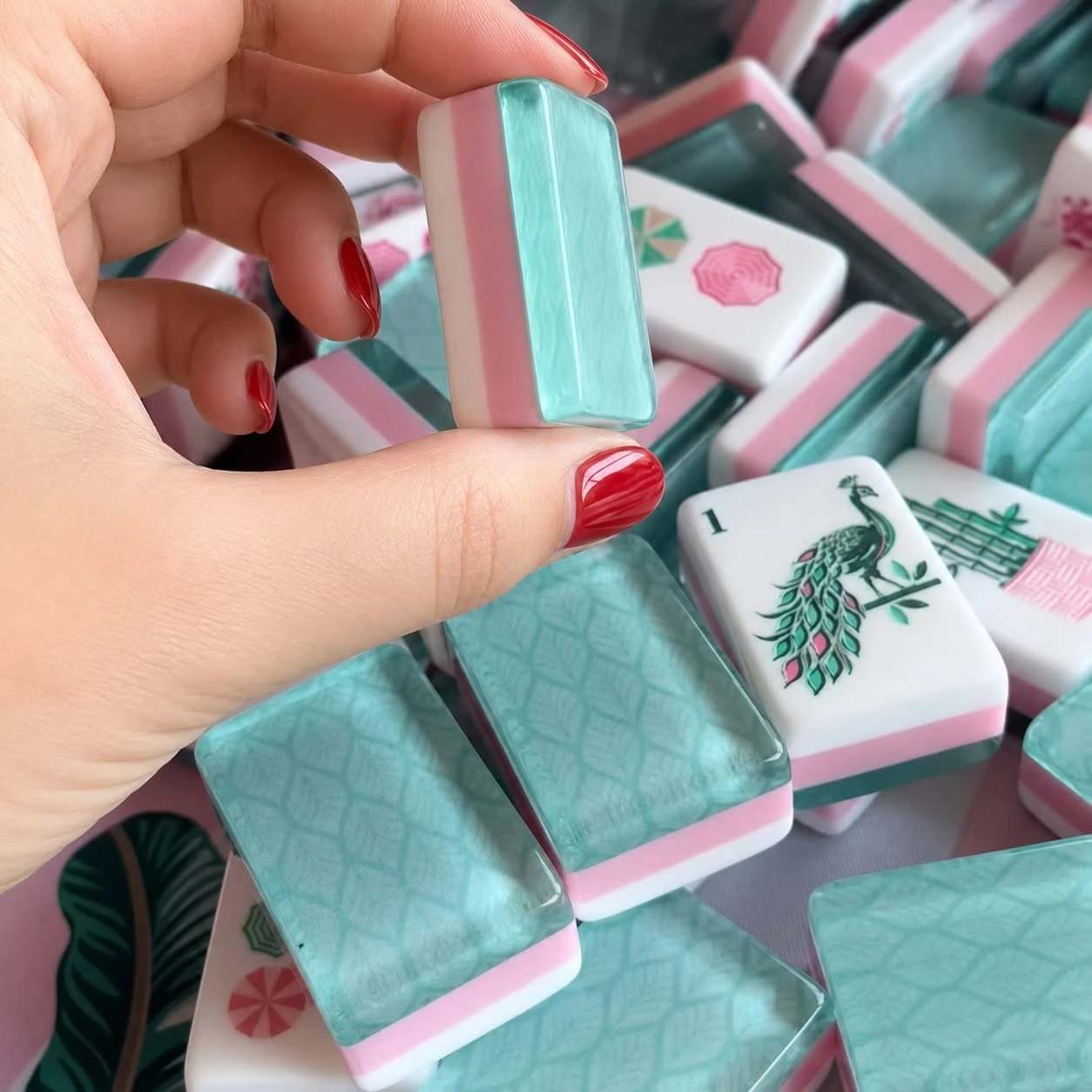 Hand holding a mahjong tile showing an aqua patterned back and pink and white layers